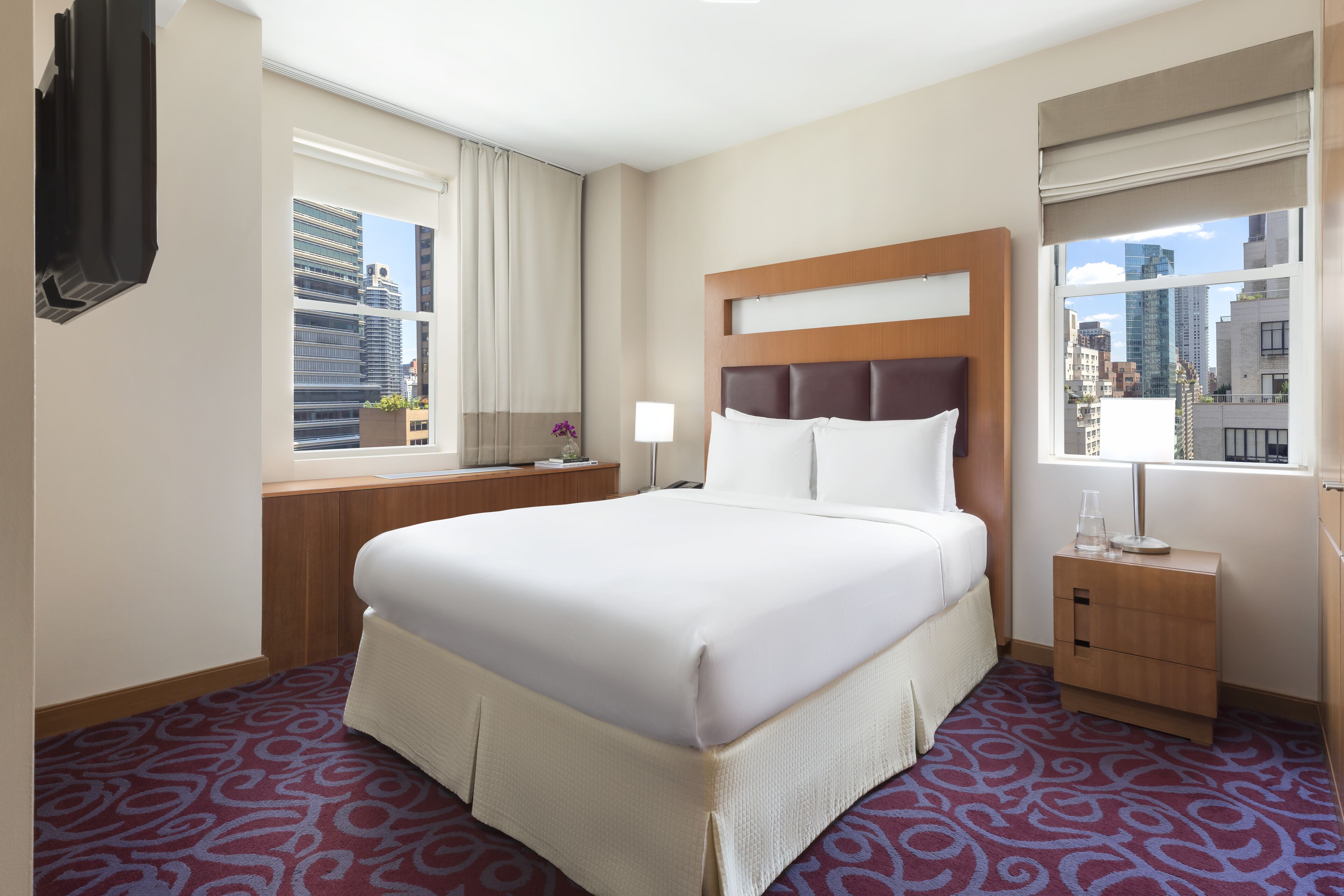 A hotel room with a large bed, a wooden nightstand, a window with a city view, and a patterned carpet.