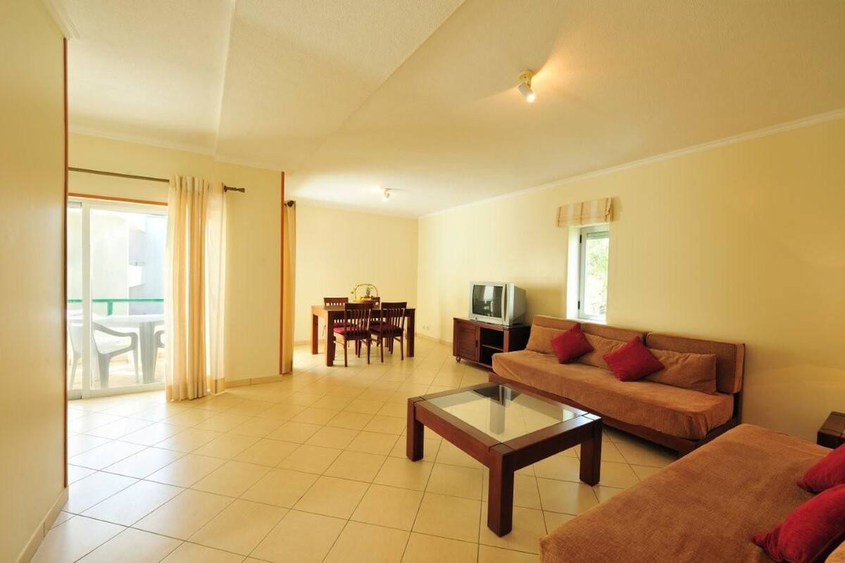 Un salon comprenant un canapé, une table basse, un téléviseur, une table à manger avec des chaises, et une porte-fenêtre coulissante donnant sur un espace extérieur.