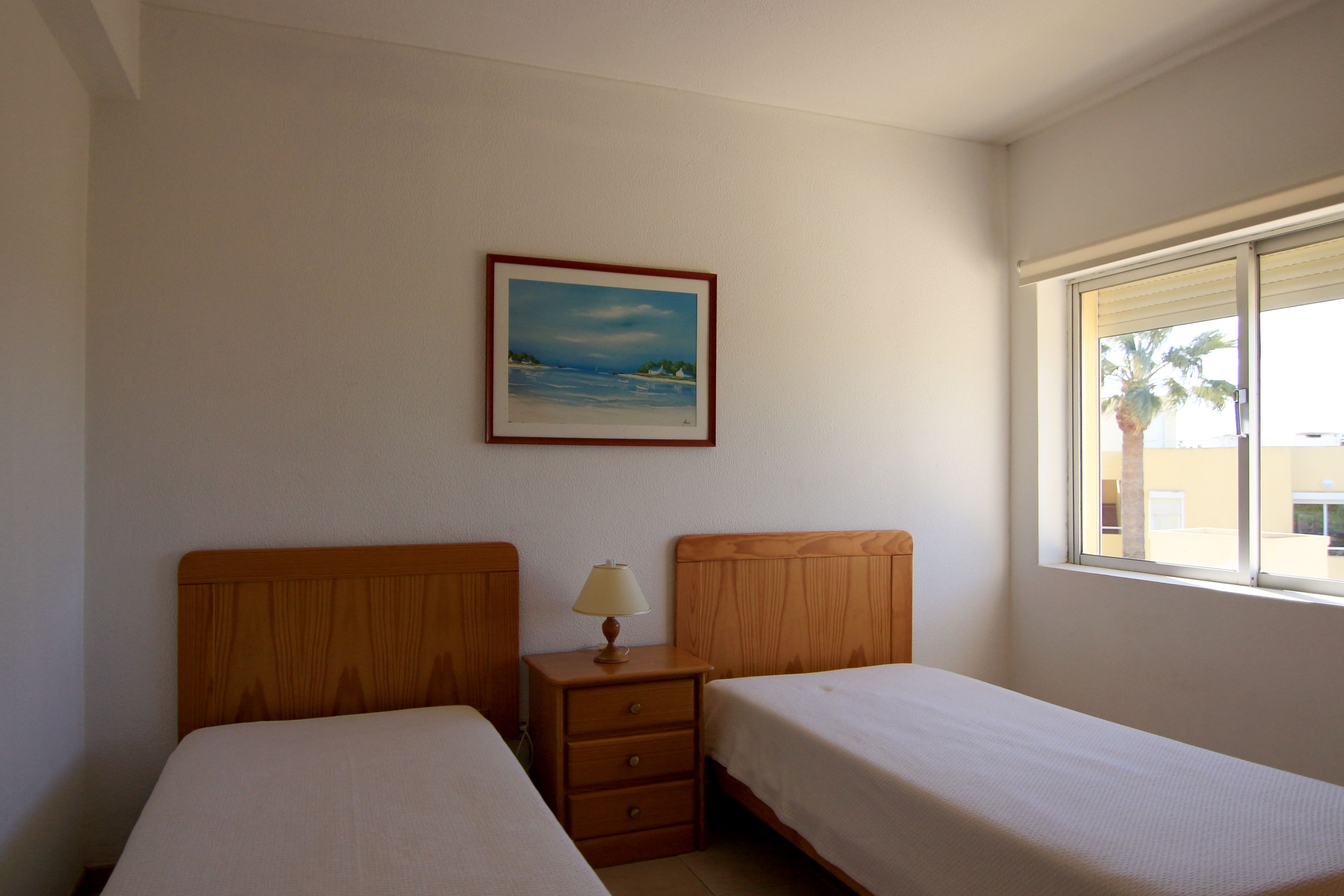 A room with two single beds, a nightstand with a lamp, a framed painting on the wall, and a window with blinds.