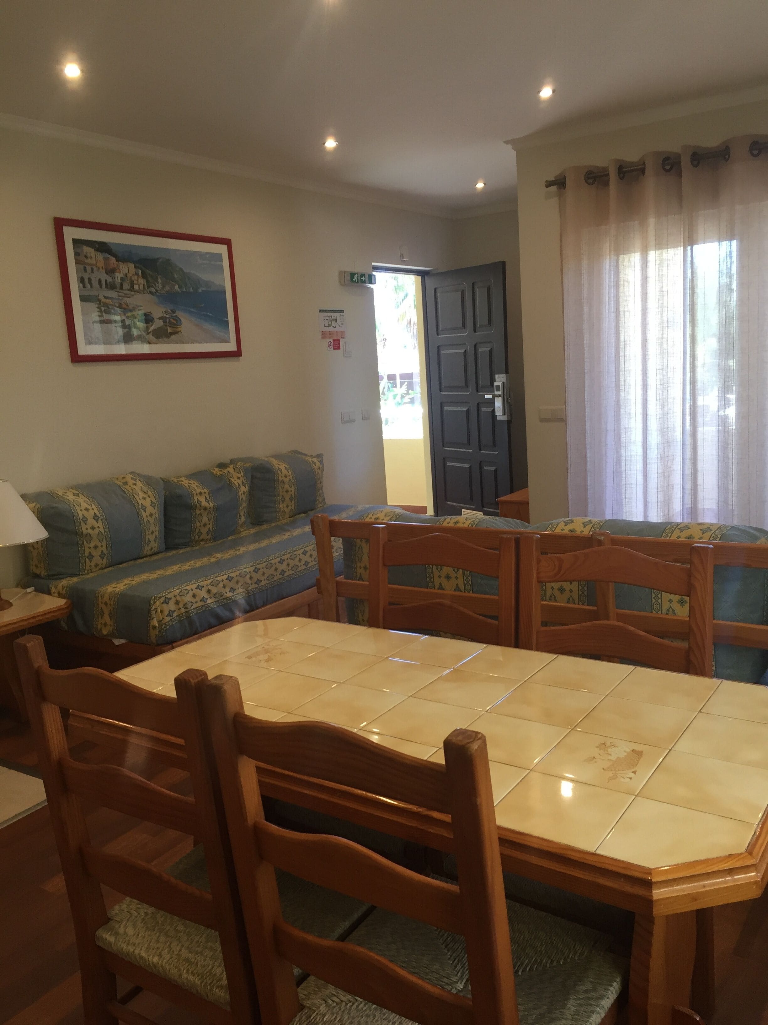 A dining area with a tiled table and wooden chairs, a sofa with patterned cushions, and a framed picture on the wall.