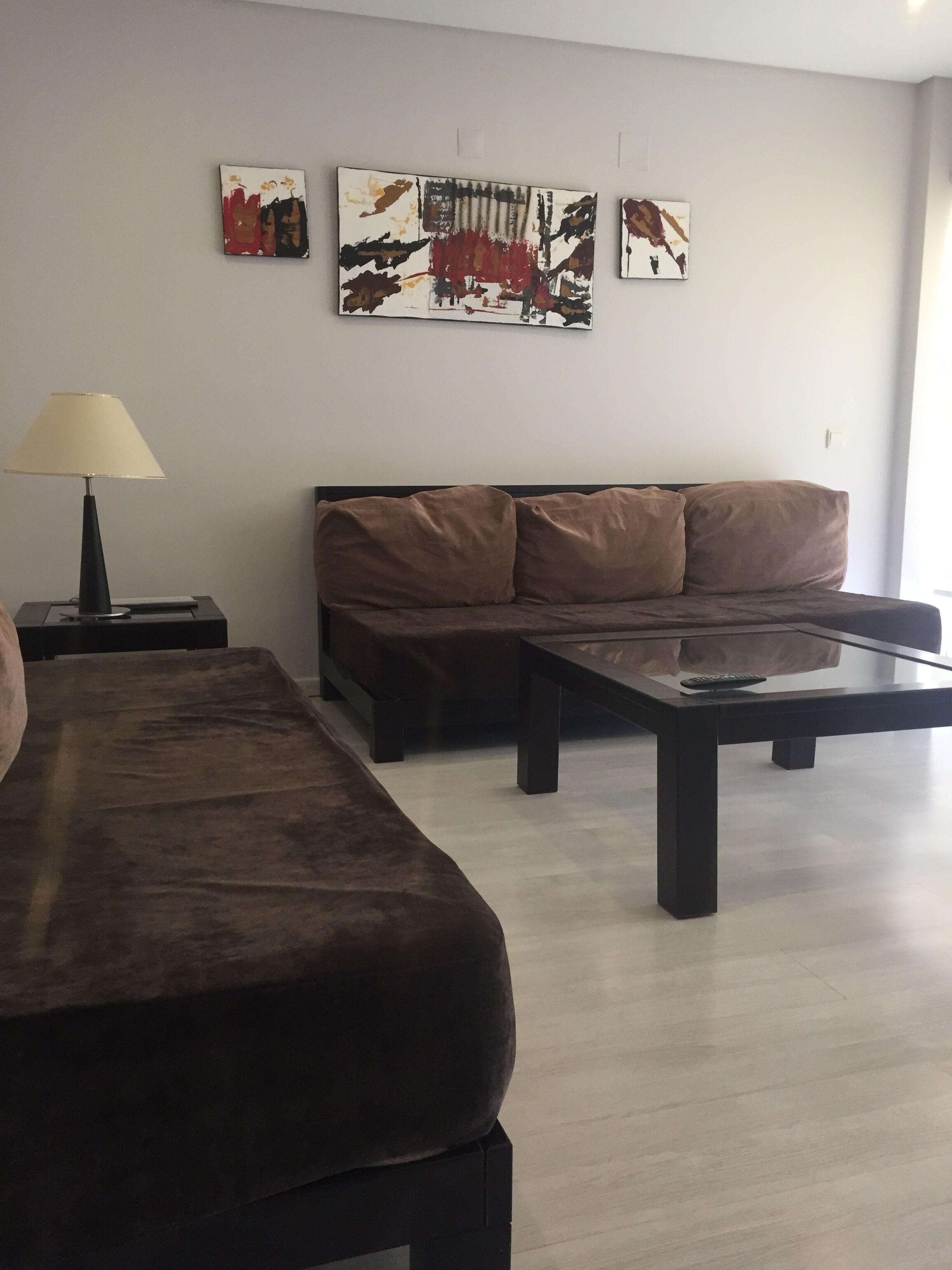 A living room with a brown sofa, a coffee table, and a lamp. There are three artworks on the wall.