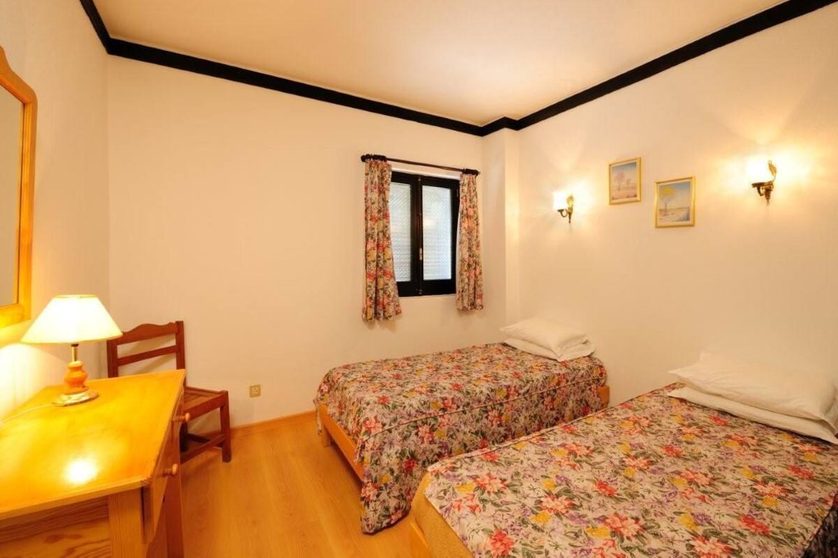 Une chambre avec deux lits simples, une fenêtre avec des rideaux à motifs floraux, une table de chevet avec une lampe et des lampes fixées au mur.