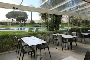 En udendørs terrasse med borde og stole, en pool og et glasomkranset område med udsigt til poolen og grøn bevoksning.