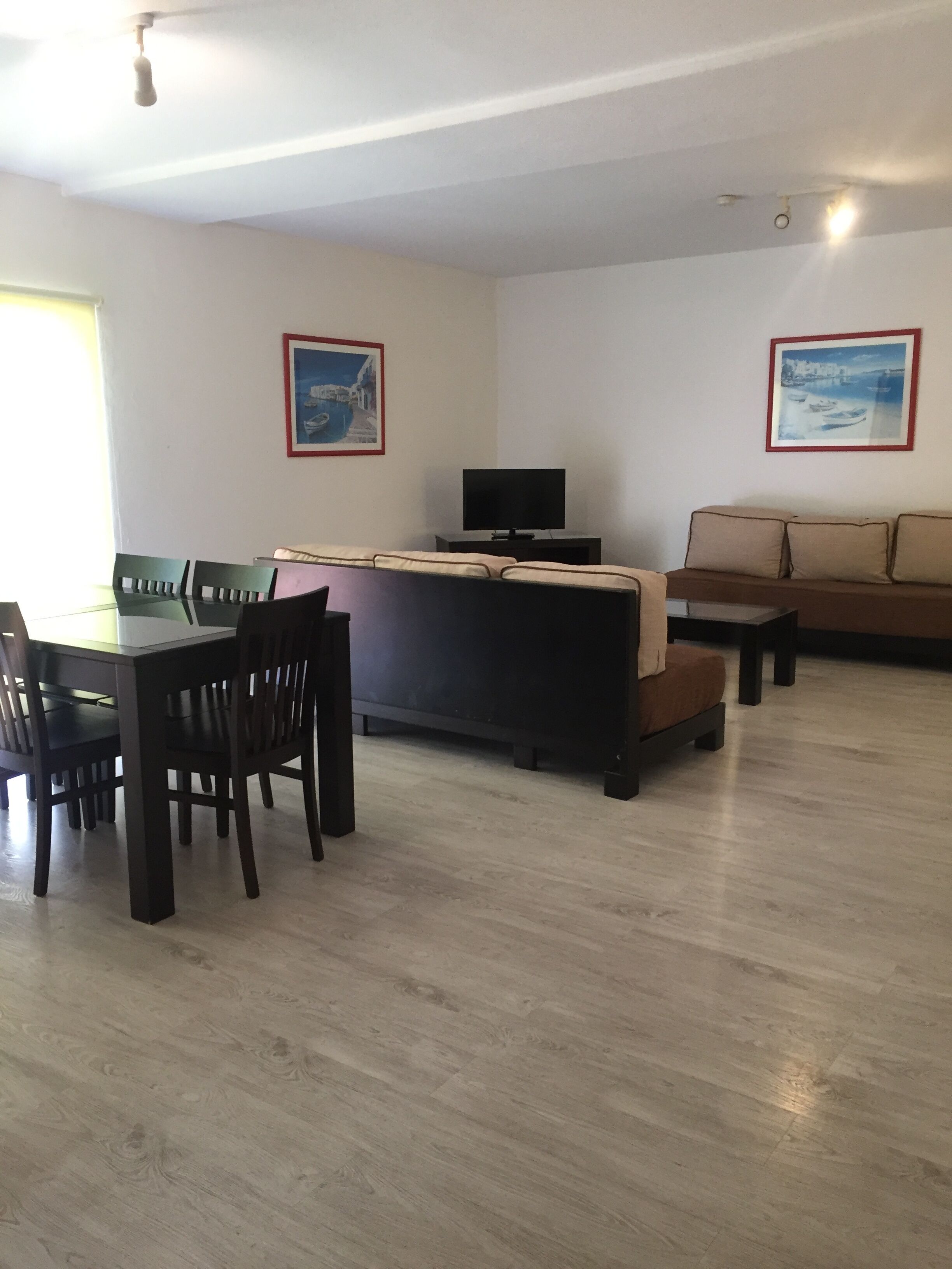 Uma sala de estar com sofá, uma mesa de jantar com cadeiras, uma televisão e duas imagens emolduradas na parede.