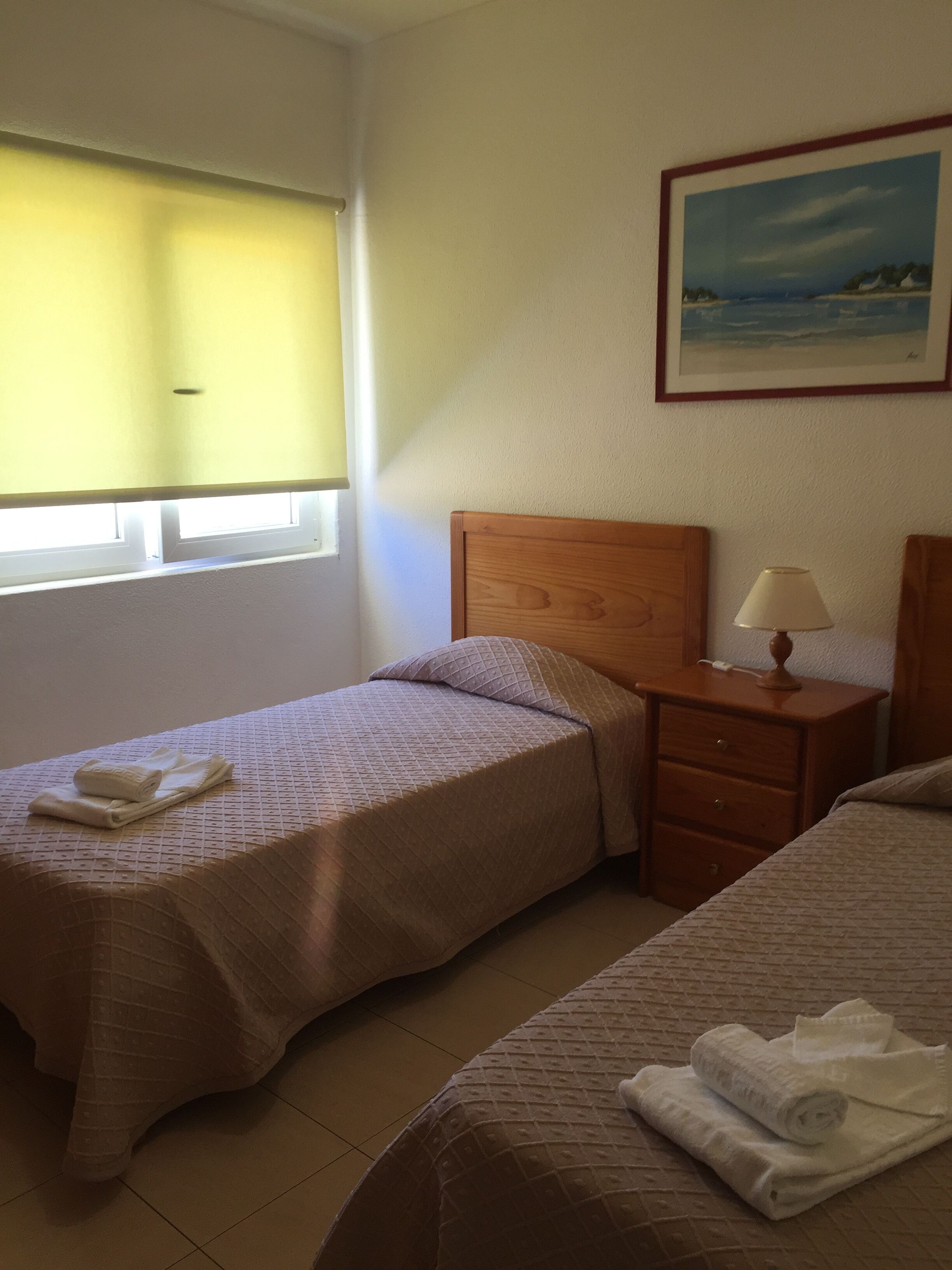 A hotel room with two beds, a nightstand, a lamp, and a painting on the wall.