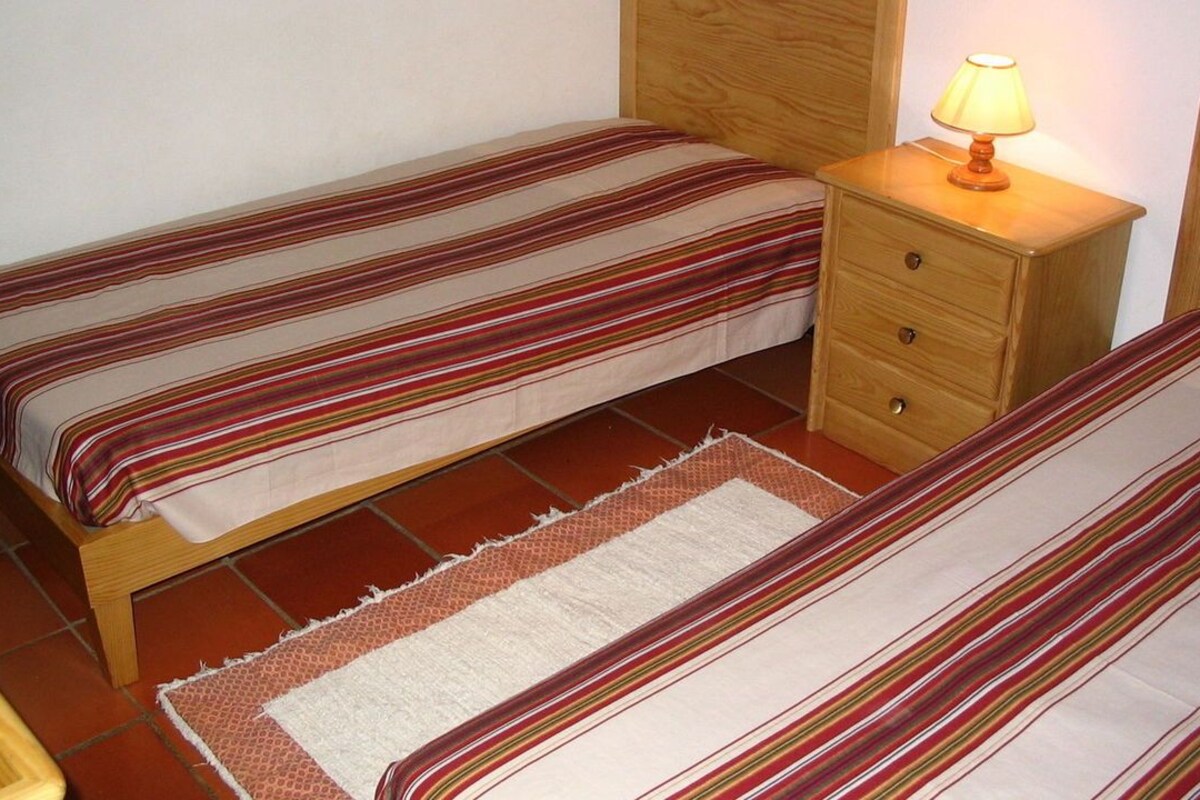 A small room with two single beds, each with striped bedding, a wooden nightstand with a lamp, and a small rug on the floor.