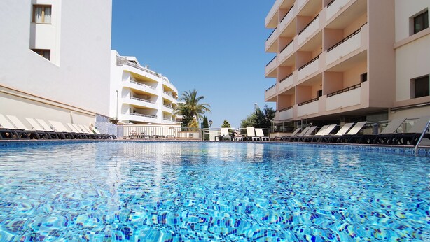 A swimming pool with clear blue water, surrounded by white buildings with balconies and lounge chairs. - Invisa Hotel La Cala (Santa Eulalia del Rio)