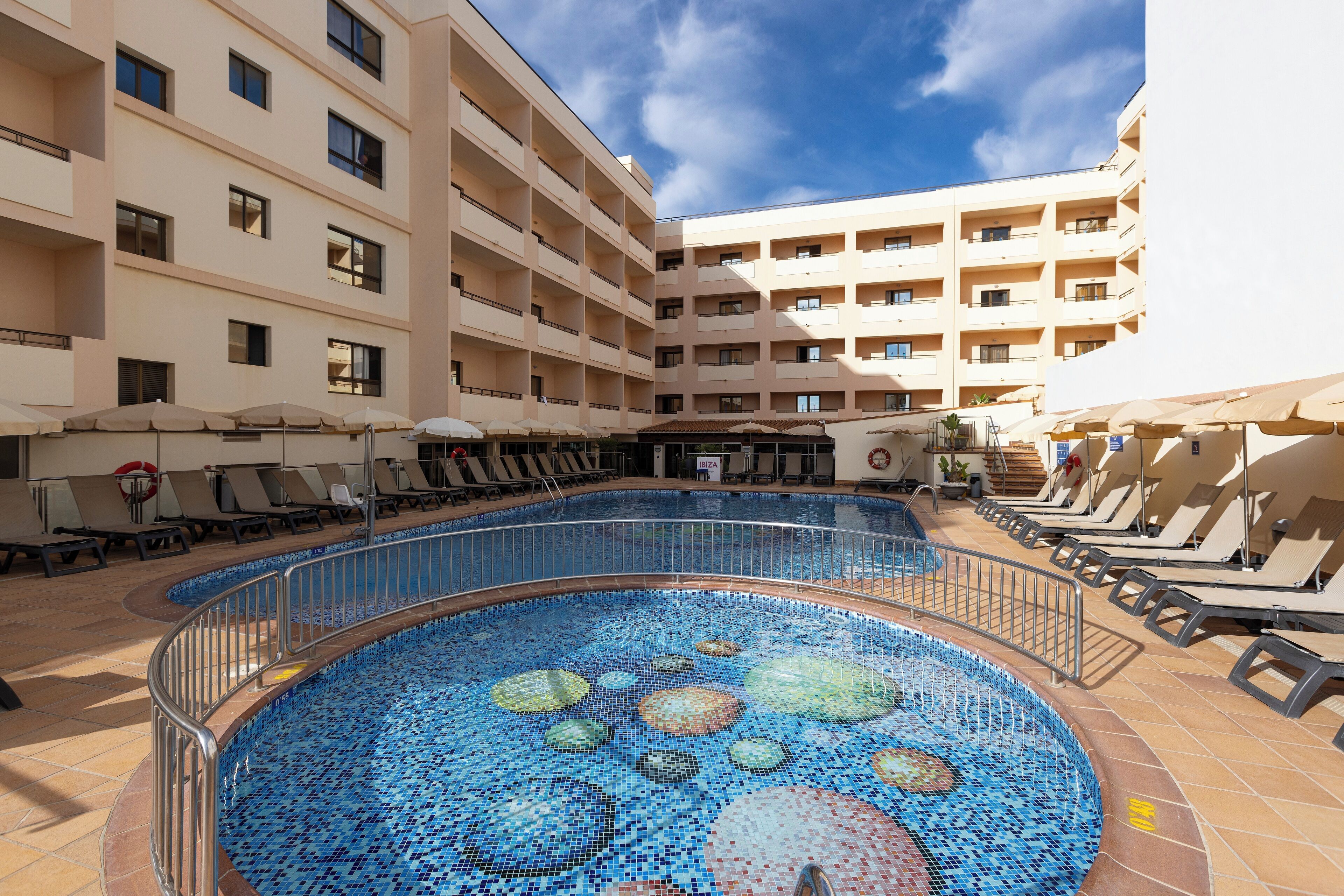 A swimming pool with a mosaic design, surrounded by lounge chairs and umbrellas, with a multi-story building in the background.