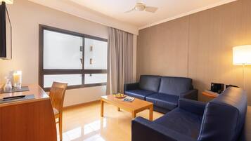 A modern hotel room with a wooden desk, a blue sofa, a coffee table, and a large window with curtains.