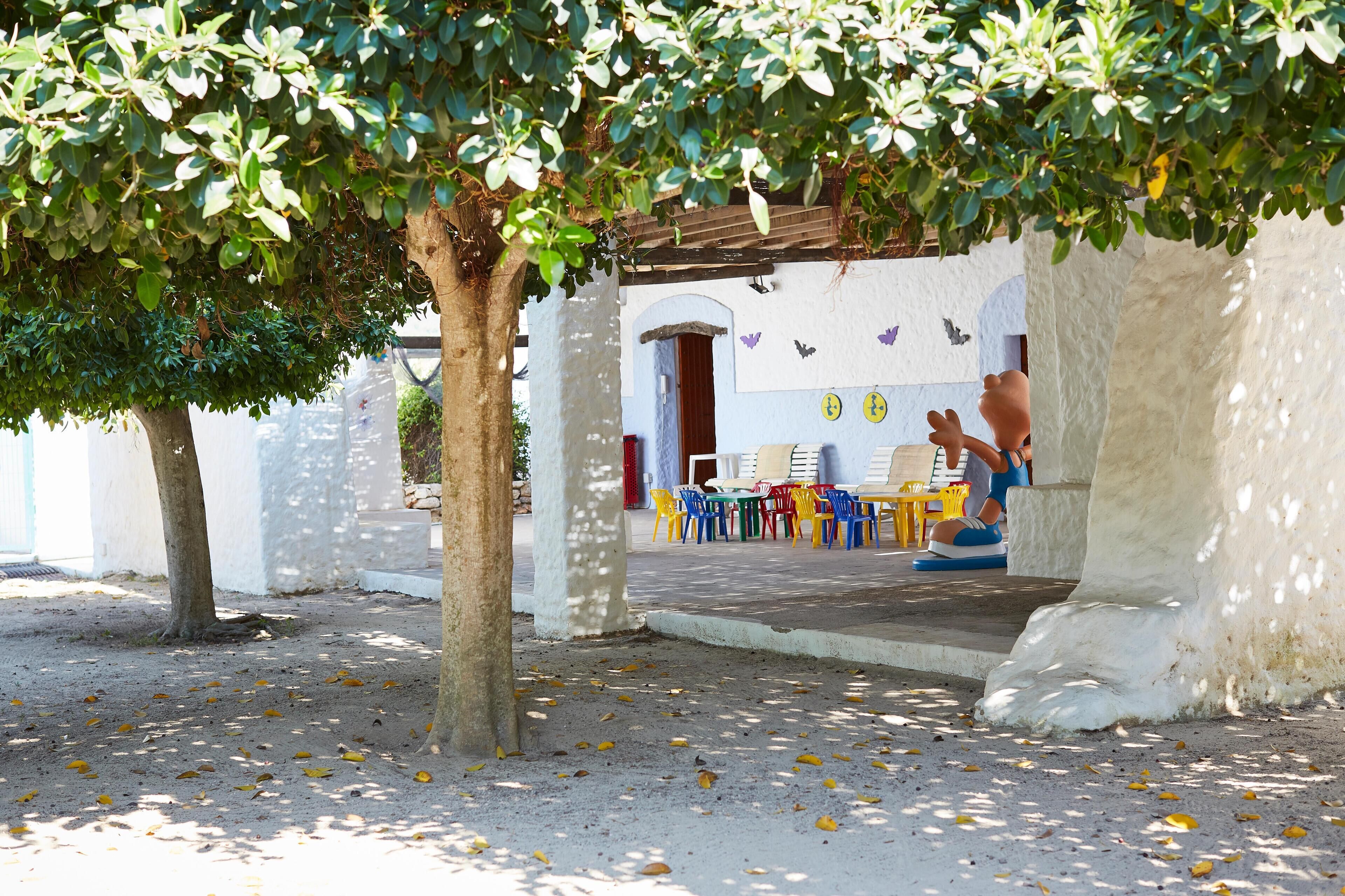 An outdoor seating area with tables and chairs, a tree, and a building with white walls and purple butterfly decorations.