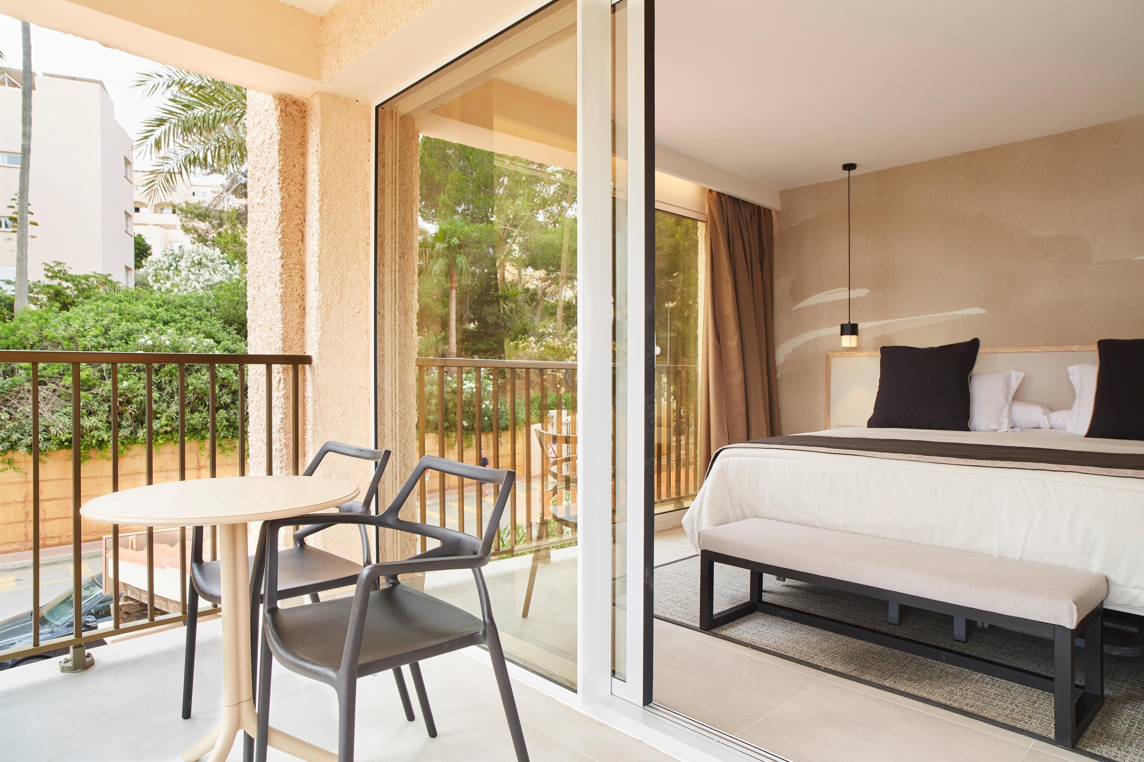 A hotel room with a bed, a bench, a table with chairs, and a balcony with a railing.