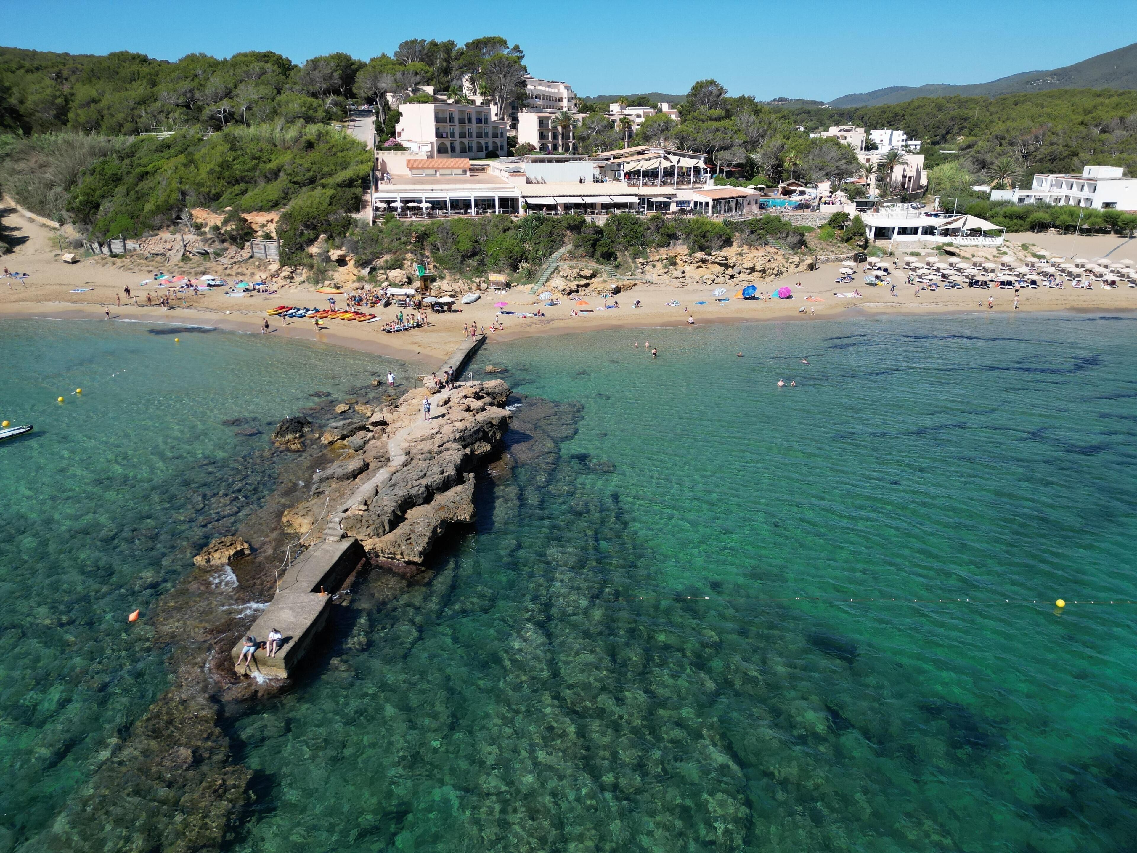 A coastal area with a rocky jetty extending into clear blue waters, surrounded by beach umbrellas and a row of buildings with balconies overlooking the sea.