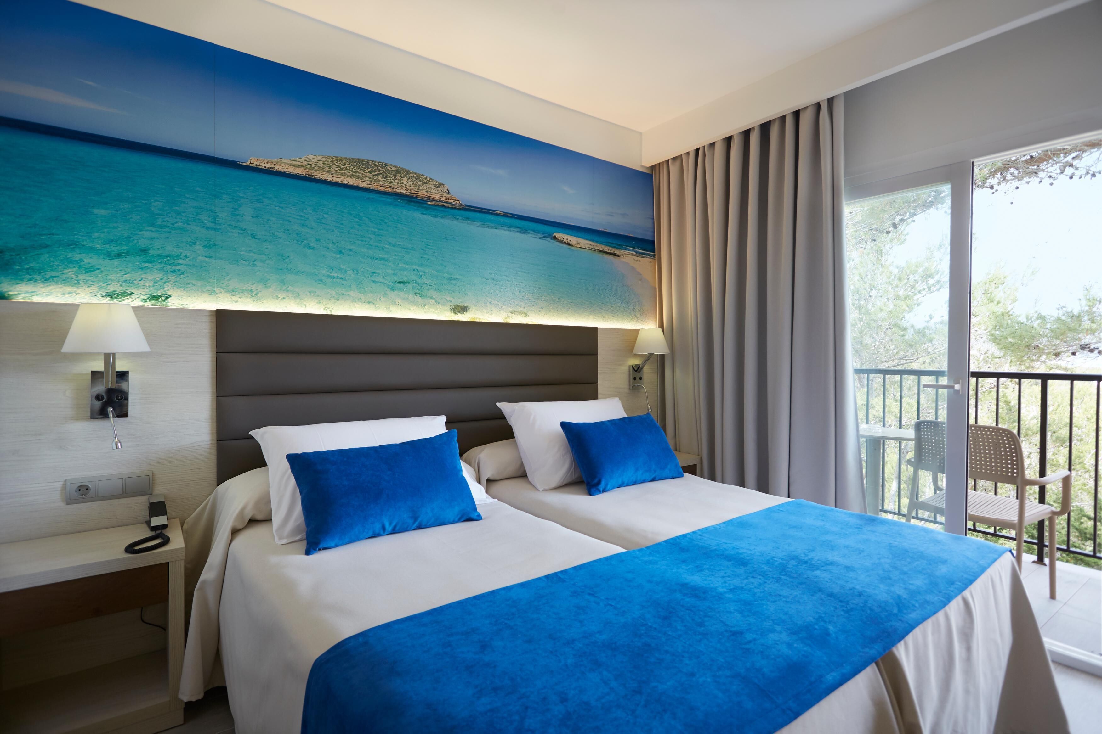 Habitación de hotel con dos camas, un ventanal con vista al mar, un balcón con sillas y un teléfono en la mesita de noche.