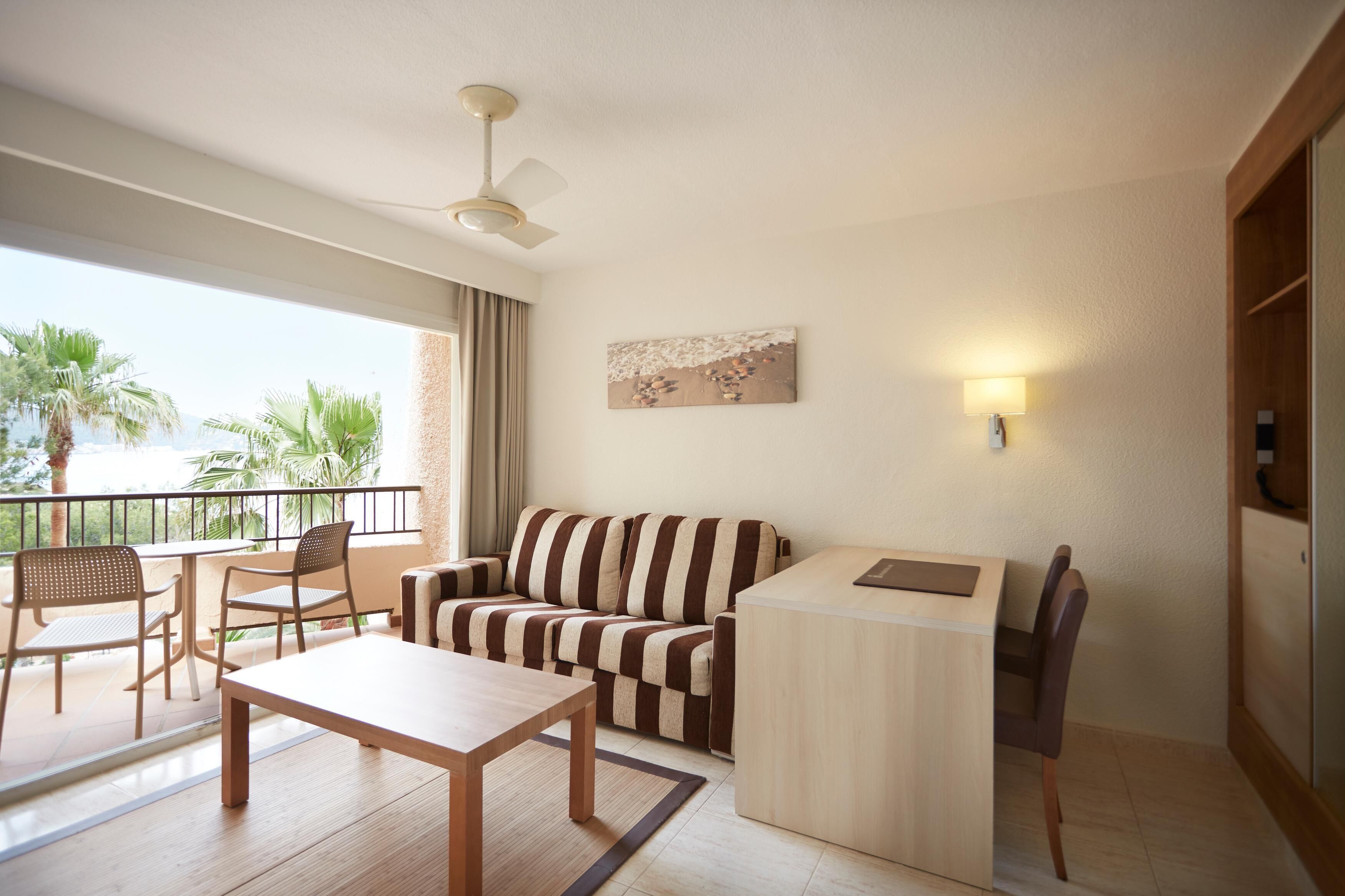 Una sala moderna con sofá, una mesa de comedor y sillas. Hay un balcón con vistas a palmeras y al mar.