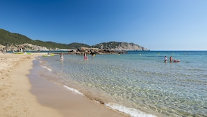 Una platja amb aigua cristal·lina, sorra daurada i turons rocosos al fons.