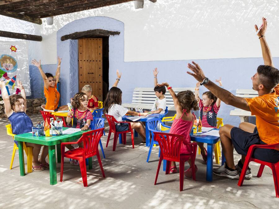 A group of children and adults seated at colorful tables in a covered outdoor area with a rustic architectural style.