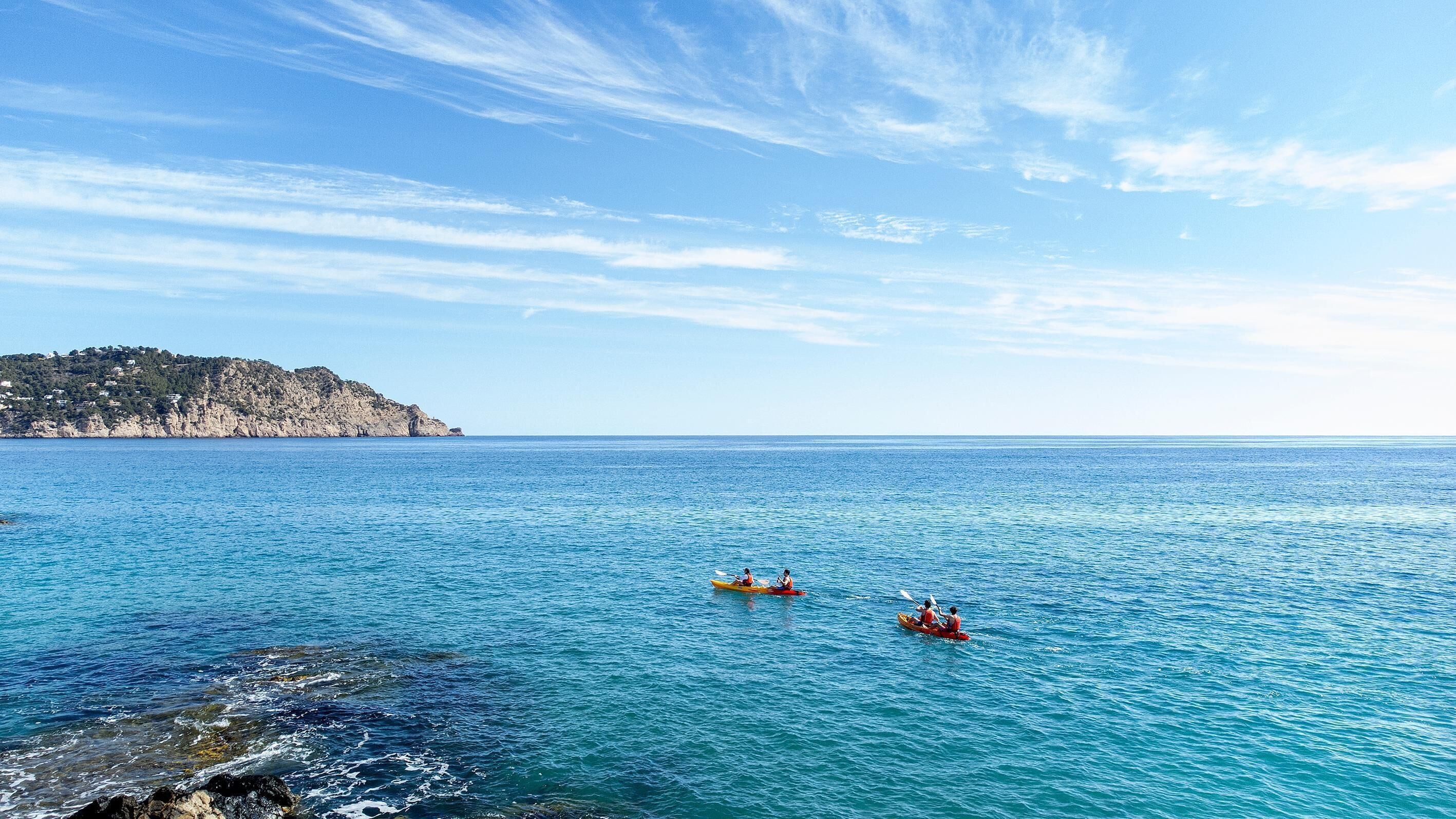 A coastal scene with calm waters, a rocky shoreline, and a distant hill with buildings.