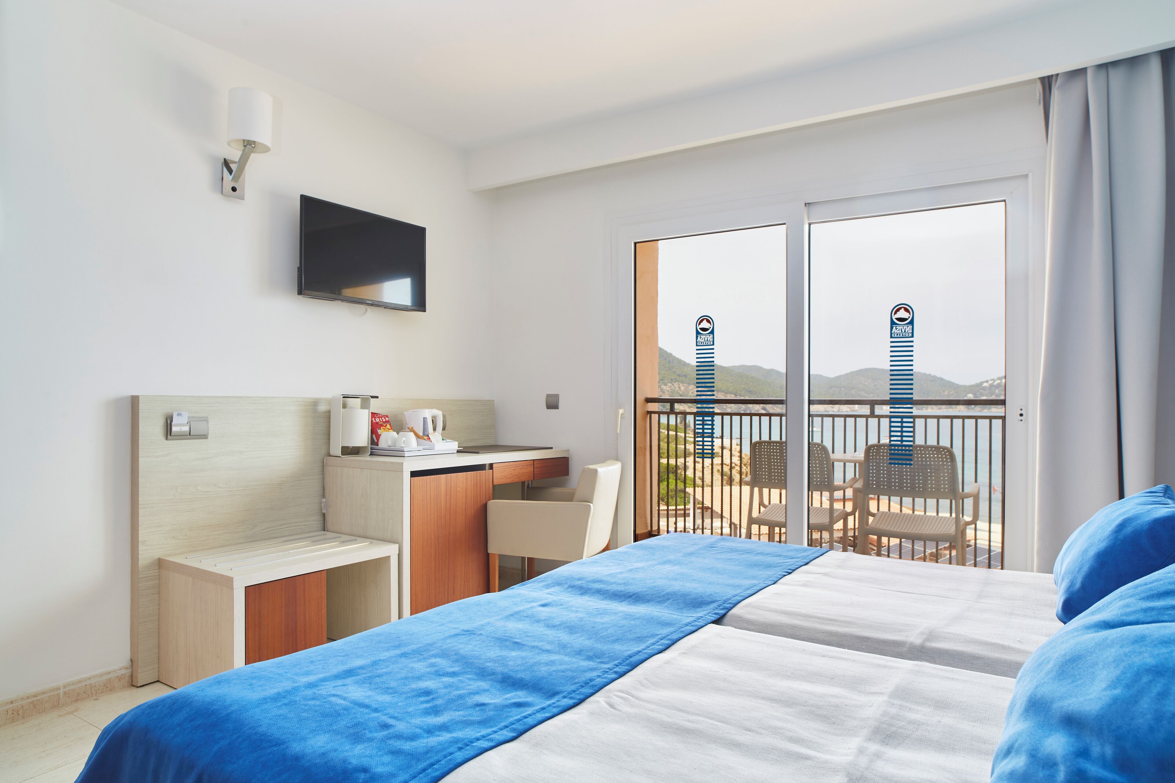 A hotel room with a bed, a desk, a chair, a TV, and a balcony with a view of mountains.