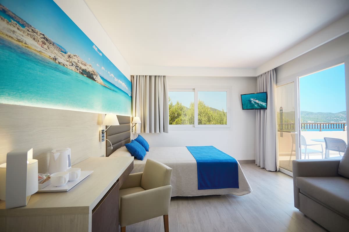 Une chambre d’hôtel avec un lit, un bureau avec un ordinateur portable, une chaise, une télévision et un balcon donnant sur la mer.