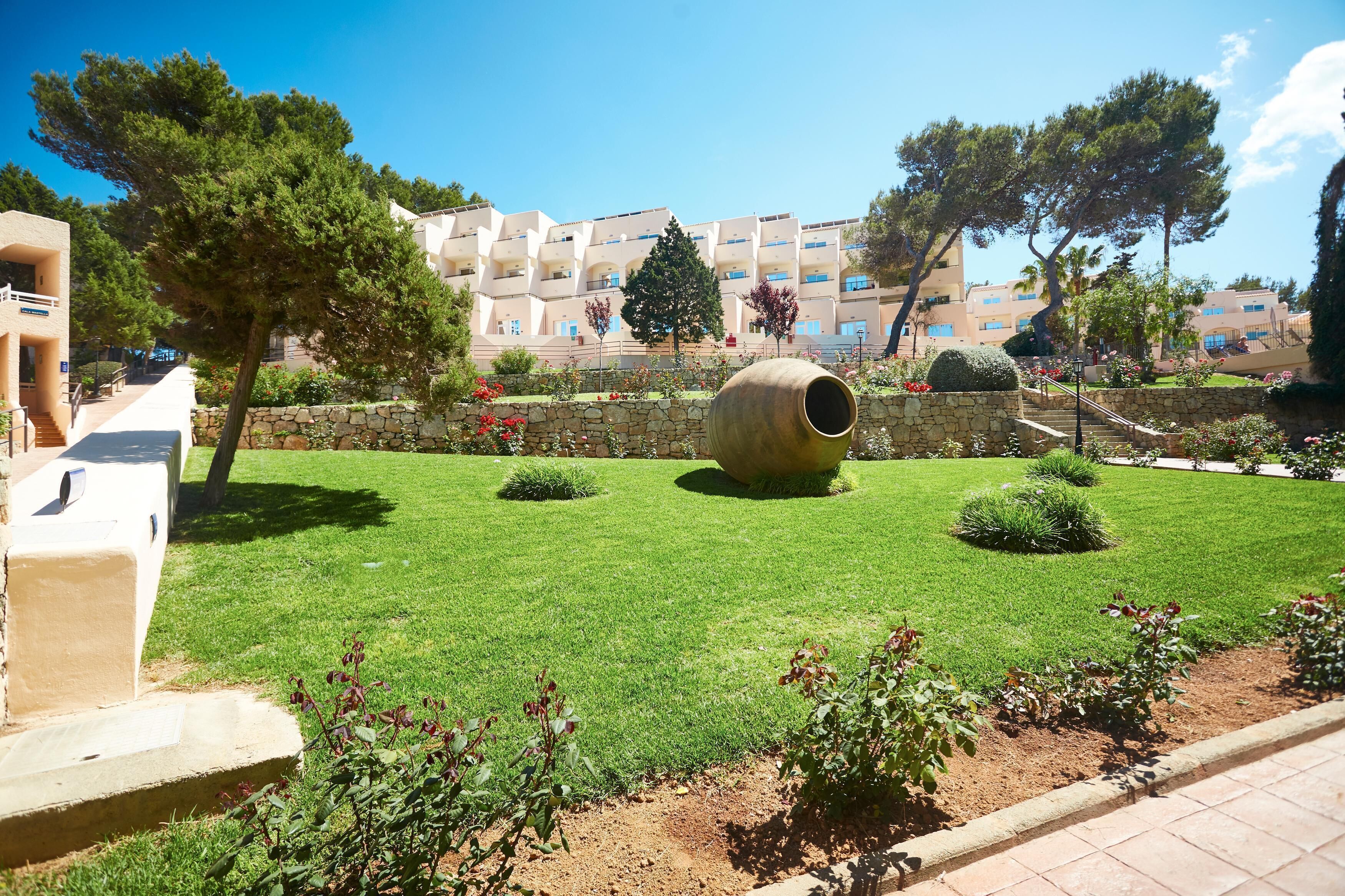 Une cour avec un mur en pierre, un grand pot cylindrique et un bâtiment avec des balcons en arrière-plan.