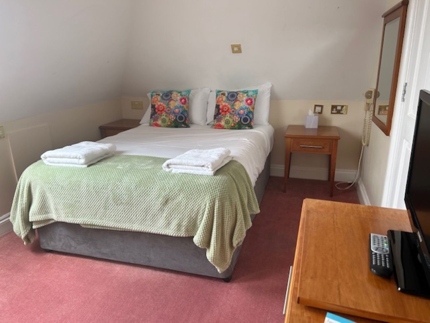 A hotel room with a bed, a bedside table with a telephone and a remote control, and a television on a wooden stand.