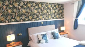 A hotel room with a bed, two bedside tables, a nightstand, a wall with a tropical wallpaper, and a window with curtains. - The Evenhill (Canterbury)