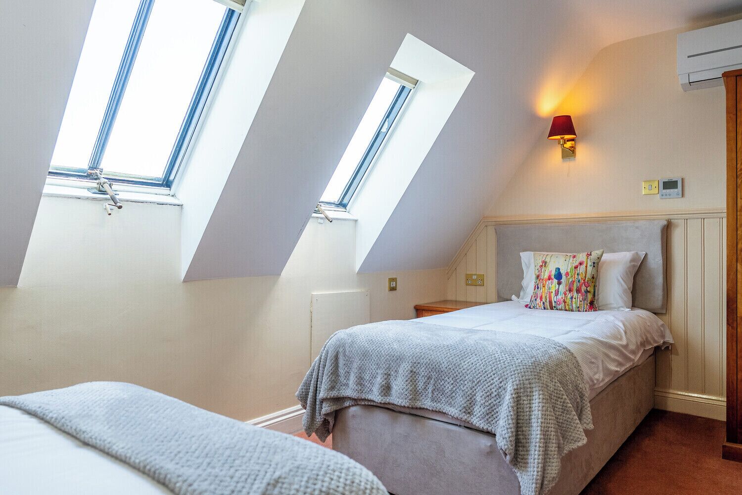 Un dormitorio con techo inclinado, tragaluces, cama con almohada colorida, mesita de noche y lámpara fijada a la pared.