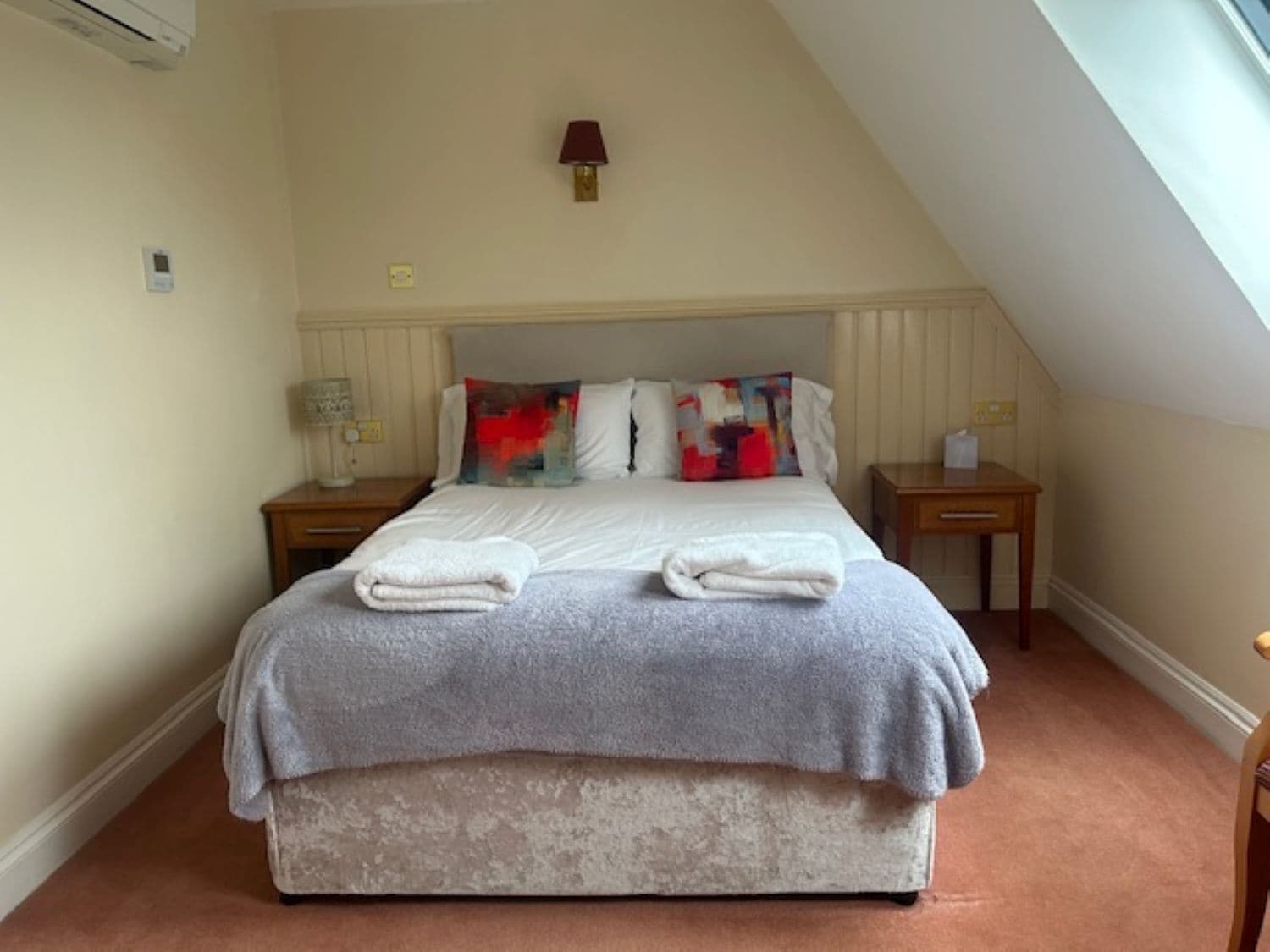 A neatly made bed with a grey blanket and two folded white towels, a bedside table with a lamp, and a small table with a tissue box.