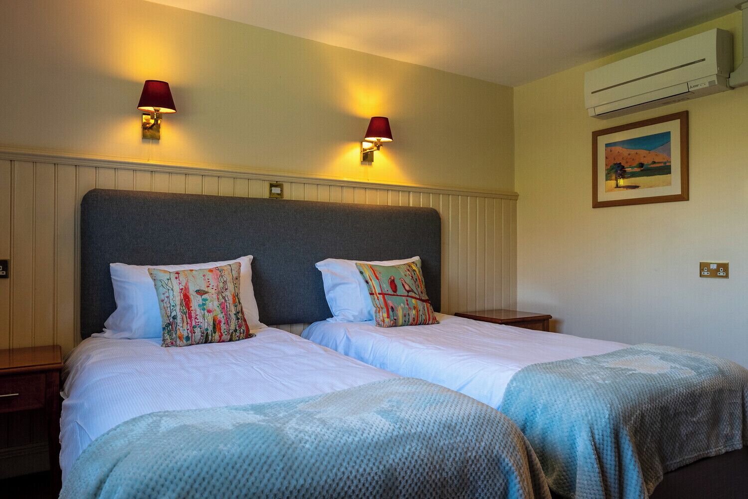 A hotel room with two beds, each with a headboard and pillows, a bedside table, an air conditioning unit, and a painting on the wall.