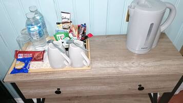 A wooden table with a white coffee maker, a water bottle, a pack of Hit chocolate biscuits, a pack of Sabicchi biscuits, and a tray of milk and tea bags.
