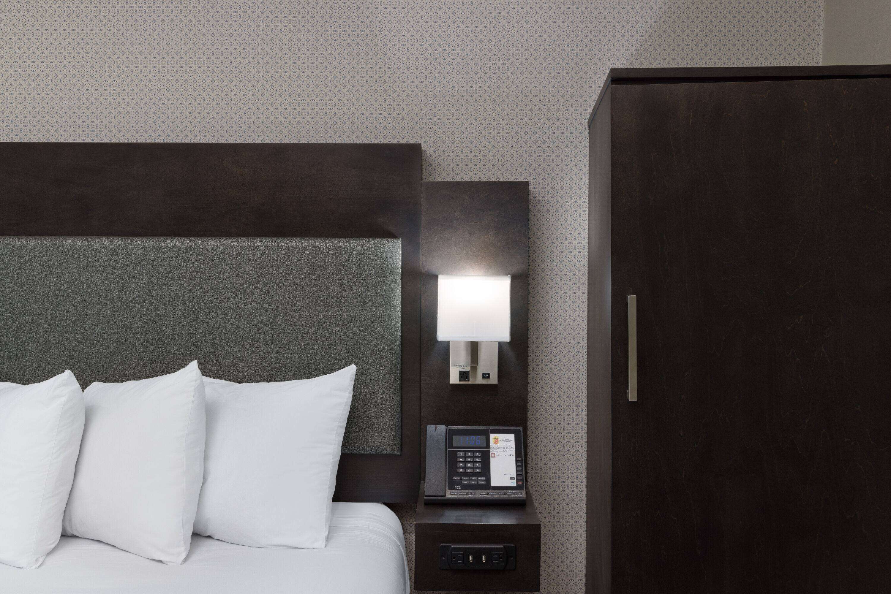 A hotel room with a bed, white pillows, a bedside table with a lamp, and a dark wooden wardrobe.