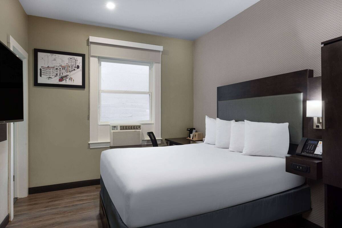 A hotel room with a large bed, a desk, a chair, a TV, and a window with a cityscape view.