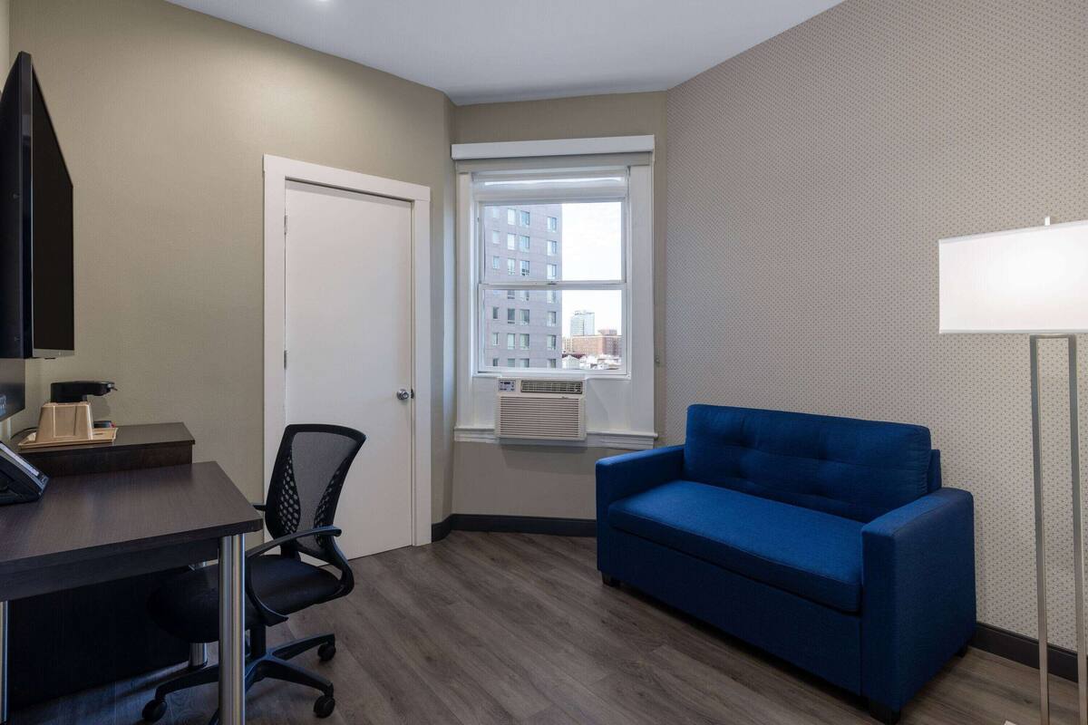 A hotel room with a desk, chair, blue sofa, and a large window with a city view.