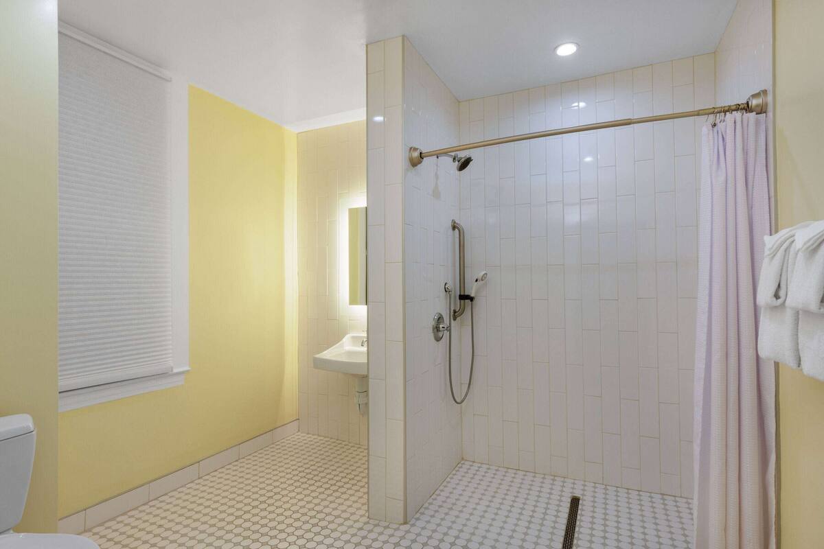 A bathroom with a shower, toilet, sink, and a window with blinds.