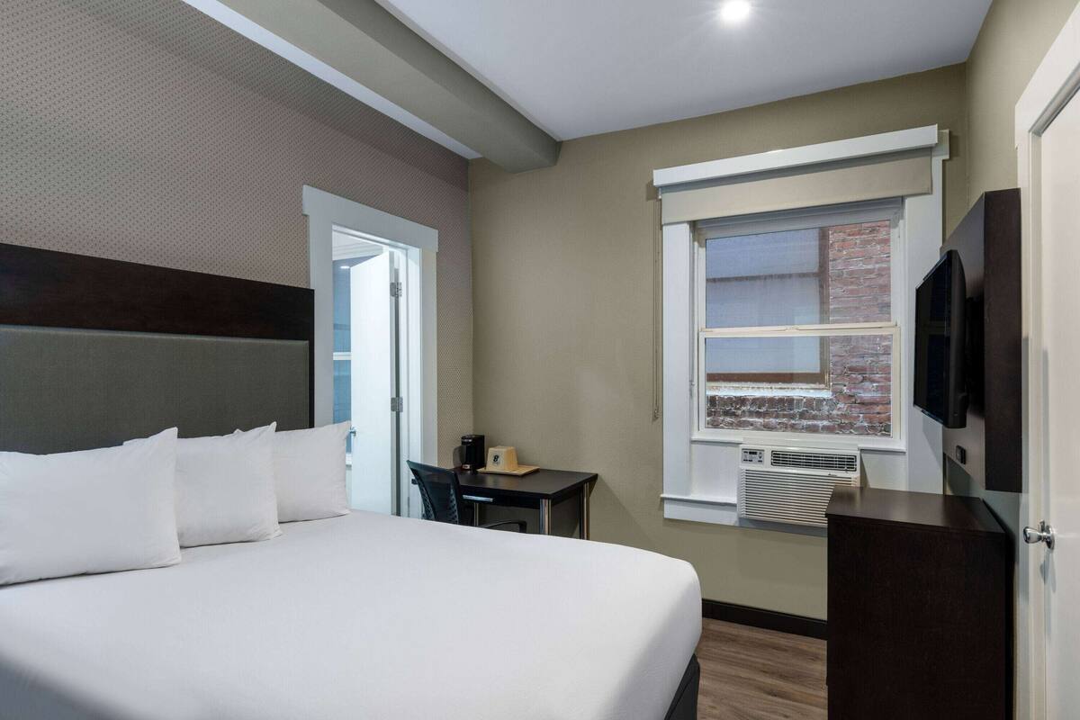 A hotel room with a large bed, a desk, a television, and a window with a view of a brick building.