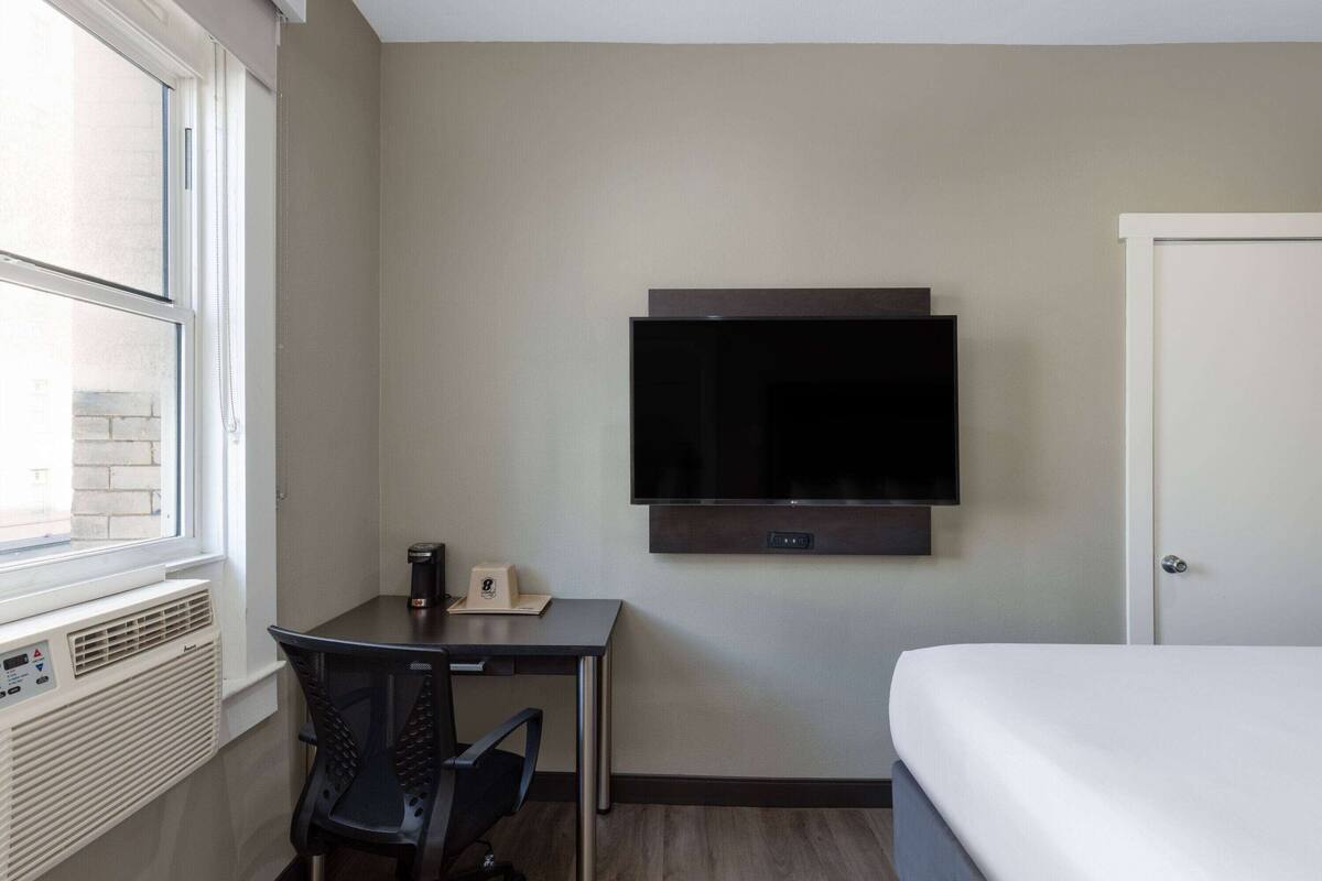 A hotel room with a bed, a desk with a chair, a television mounted on the wall, and a window.
