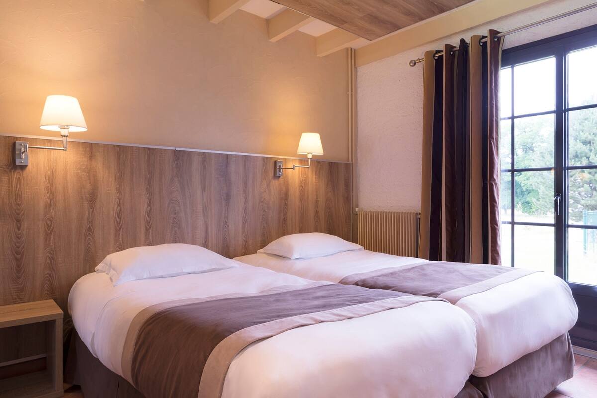 A hotel room with two beds, wooden headboard, bedside lamps, a window with curtains, and a radiator.