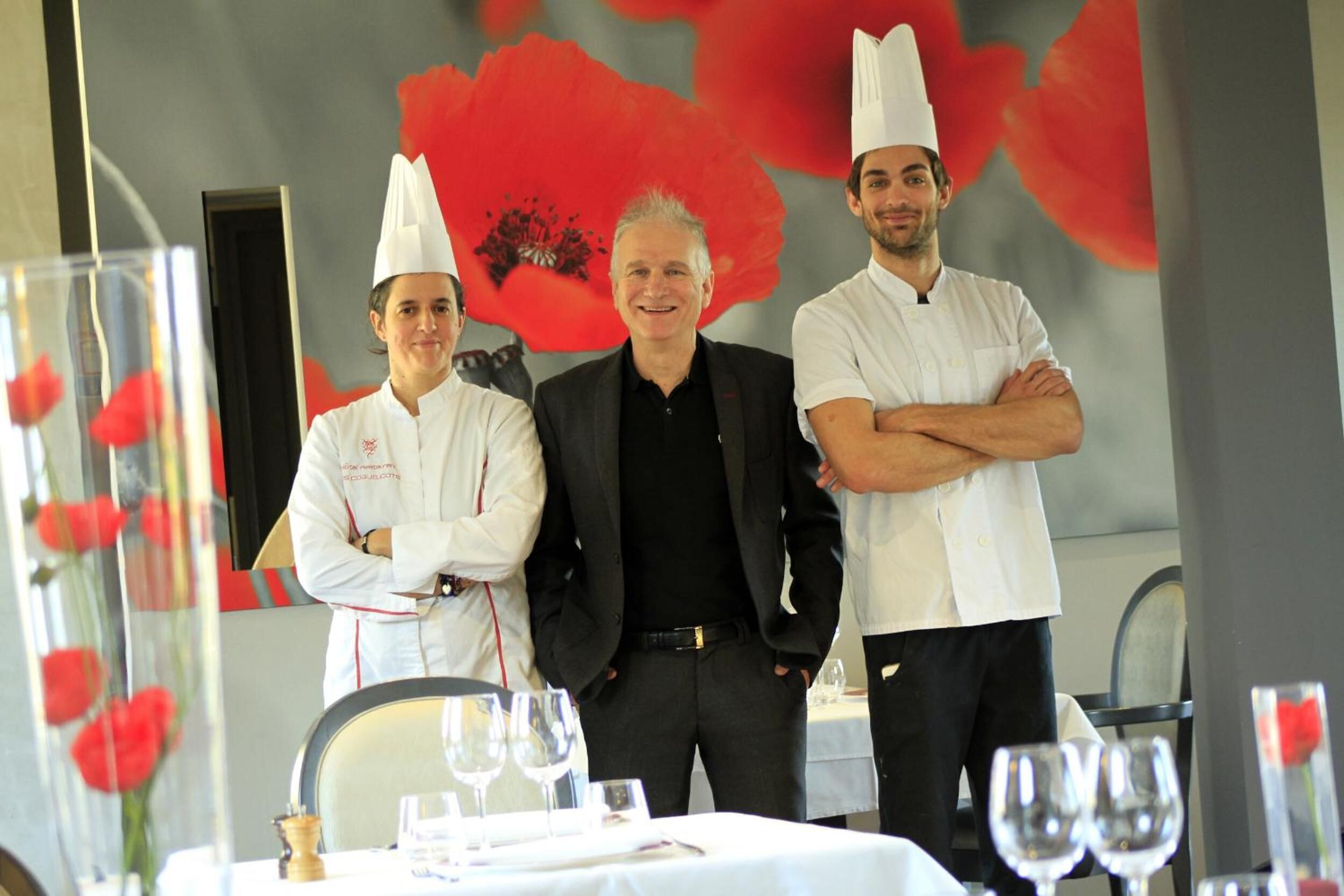 Tres personas de pie en un comedor con una mesa dispuesta para una comida, un fondo floral rojo y un espejo que refleja la escena.
