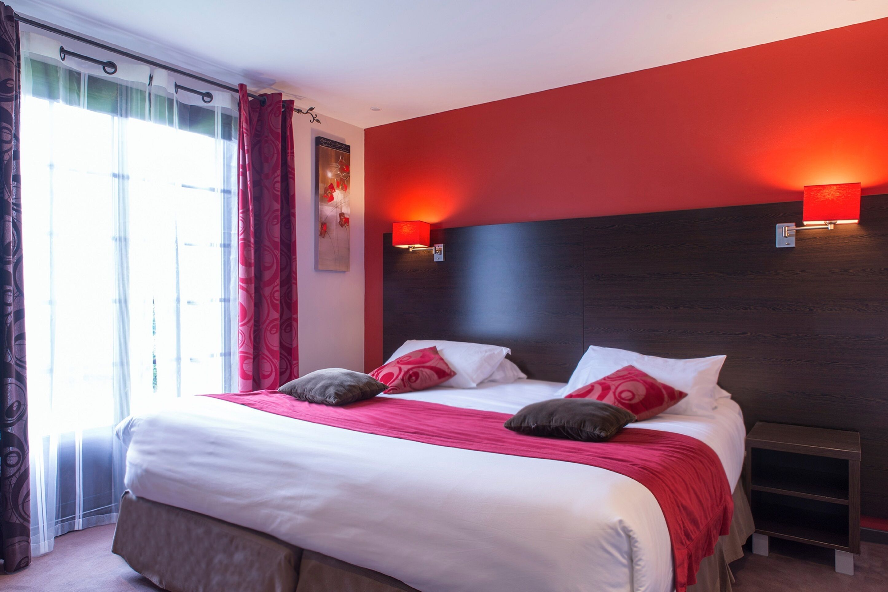 Una habitación de hotel con una cama grande, ropa de cama roja y blanca, un cabecero oscuro y una mesita de noche.