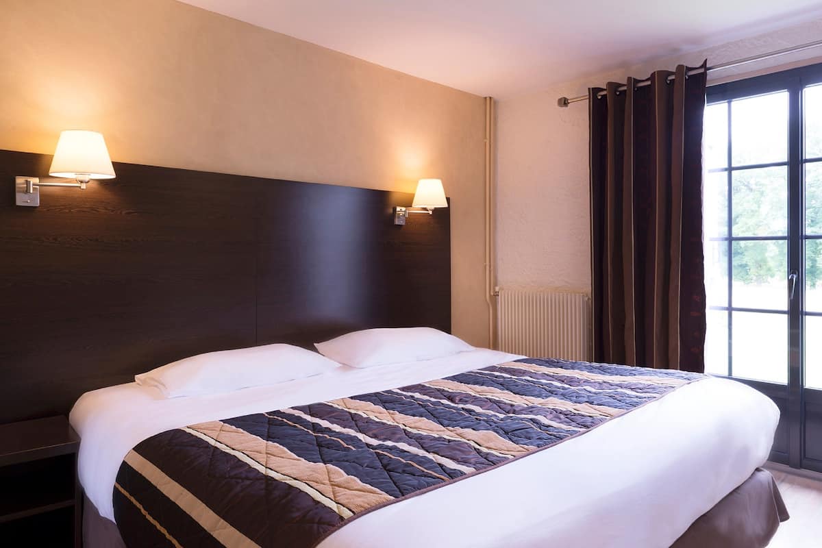 A hotel room with a large bed, two wall-mounted lamps, a window with curtains, and a radiator.