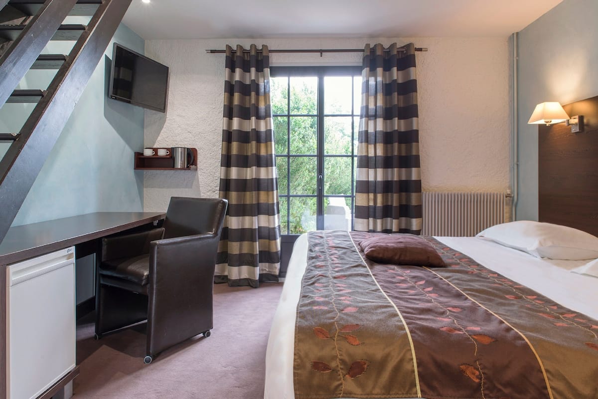 A bedroom with a bed, a desk, a chair, a TV, and a window with curtains.