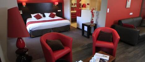 Habitación de hotel con una cama, dos sillones rojos, una mesita de centro con una tetera y tazas, y una pequeña mesa con una revista.