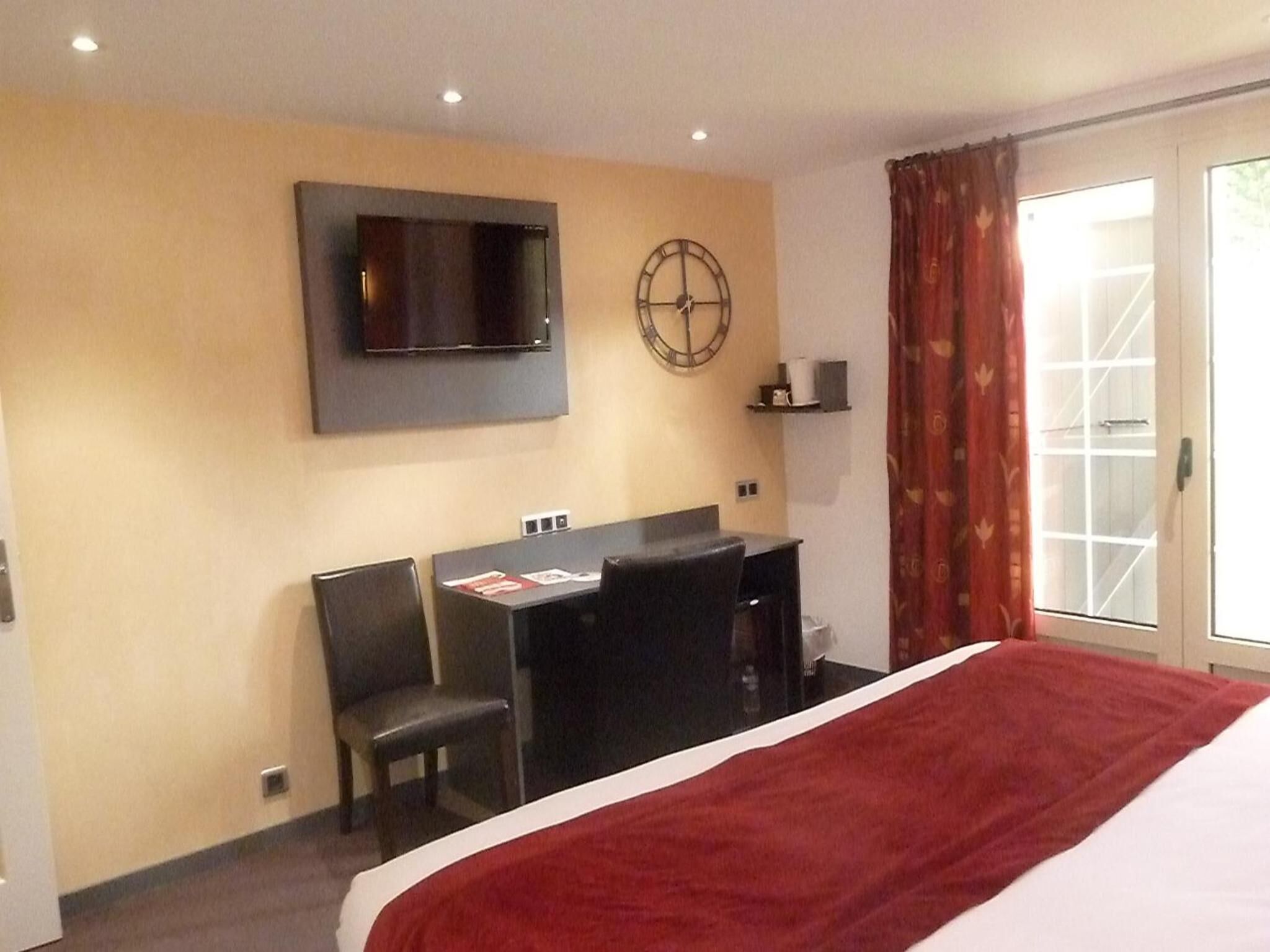 A hotel room with a bed, a desk, two chairs, a TV mounted on the wall, a clock, and a window with curtains.