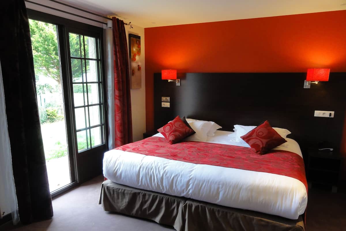 A hotel room with a large bed, red and white bedding, a view of greenery through a window, and a red accent wall.