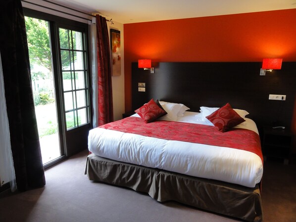 Habitación de hotel con una cama grande, ropa de cama roja y blanca, vistas a zonas verdes por una ventana y una pared de acento roja.
