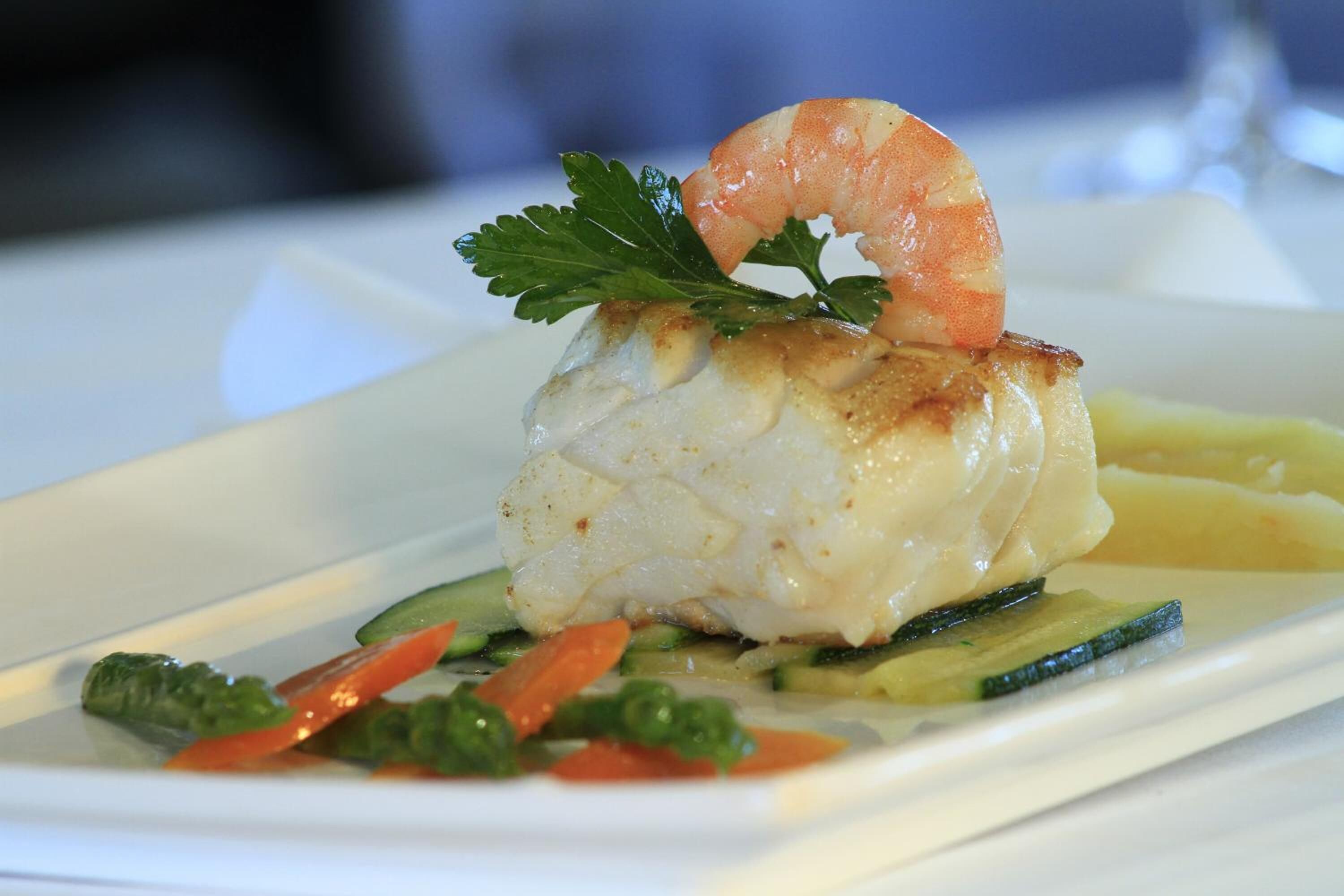 Un plato con un trozo de pescado blanco, un camarón y una guarnición de perejil, acompañado de vegetales en rodajas.