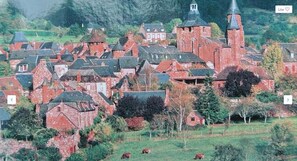 A historic village with traditional red brick buildings, a prominent church with a tall spire, and a pastoral setting with grazing cows. - The Originals Relais, Hôtel Restaurant Les Coquelicots (Saint-Pardoux-l'Ortigier)