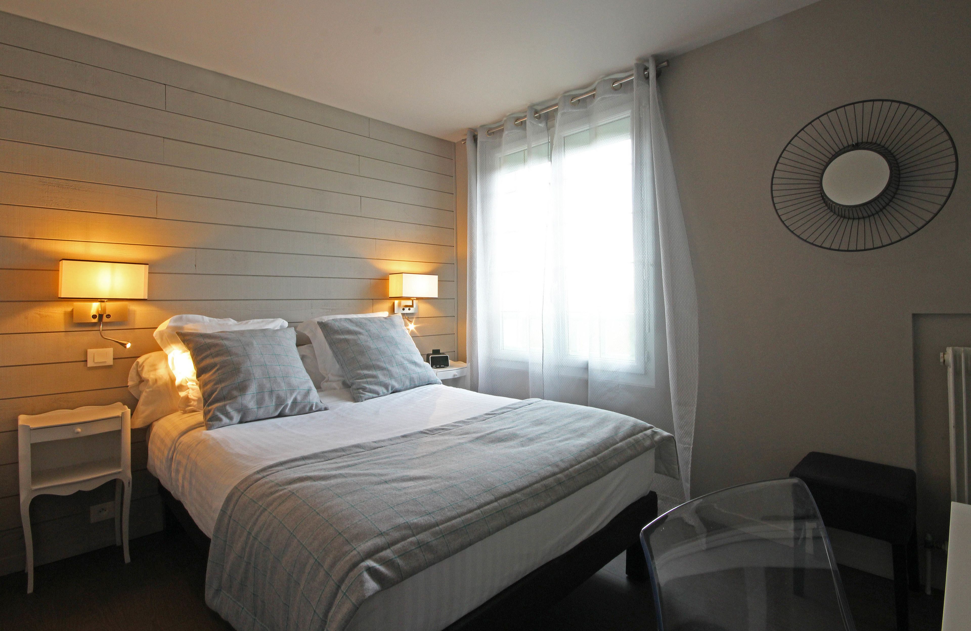 Ein Schlafzimmer mit einem Bett, zwei Nachttischen mit Lampen, einem Sessel, einem runden Spiegel an der Wand und einem Fenster mit Vorhängen.