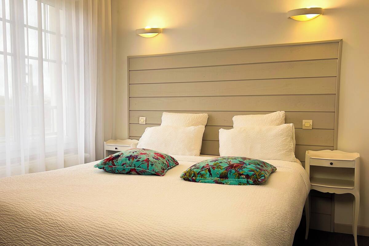 Un lit bien fait, avec du linge de lit blanc et deux coussins décoratifs, une tête de lit en bois, des tables de chevet avec des lampes et des rideaux légers.