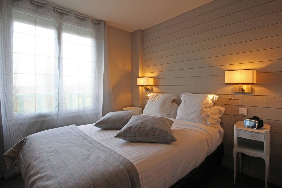Un lit bien fait avec deux oreillers, une table de chevet avec une lampe et une petite table de nuit avec une horloge.